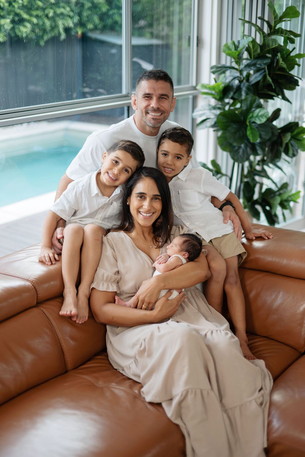 Happy family of five posing on a brown leather sofa, with parents, two boys, and a newborn baby near a pool.