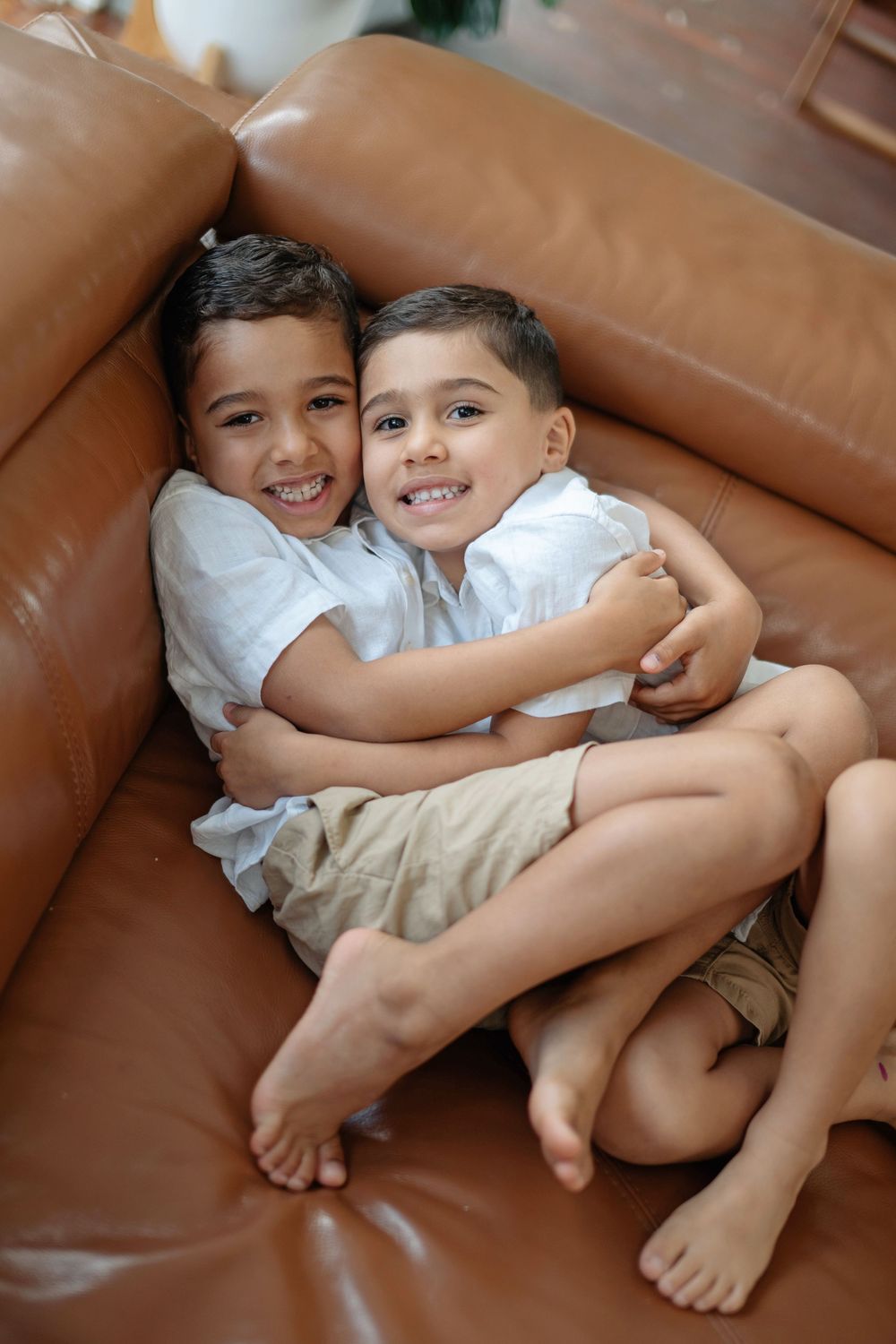 Two young brothers hugging and smiling on a brown leather sofa, wearing white shirts and barefoot.
