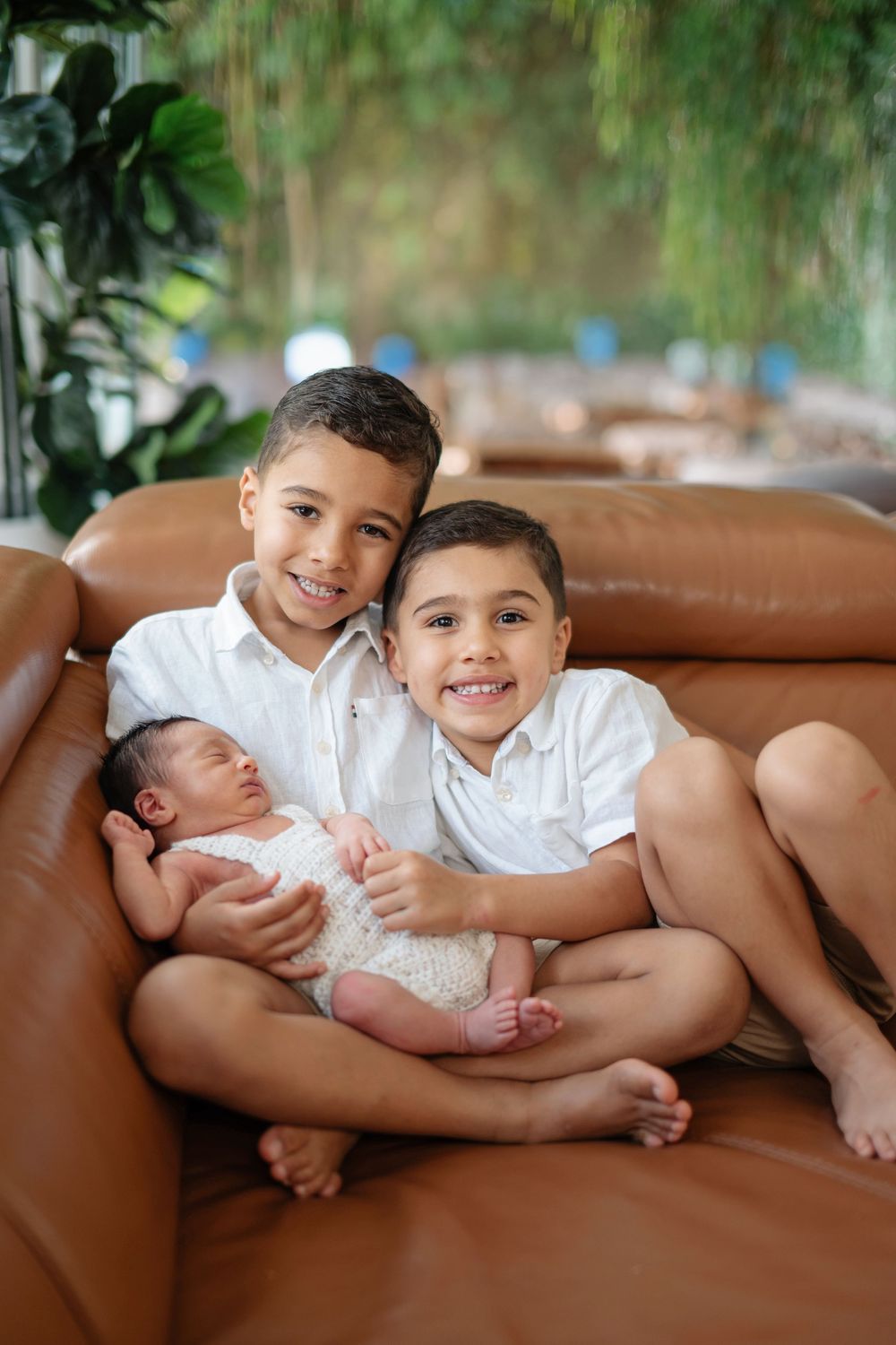 Two smiling boys in white shirts sit on a brown leather sofa holding a sleeping newborn baby.
