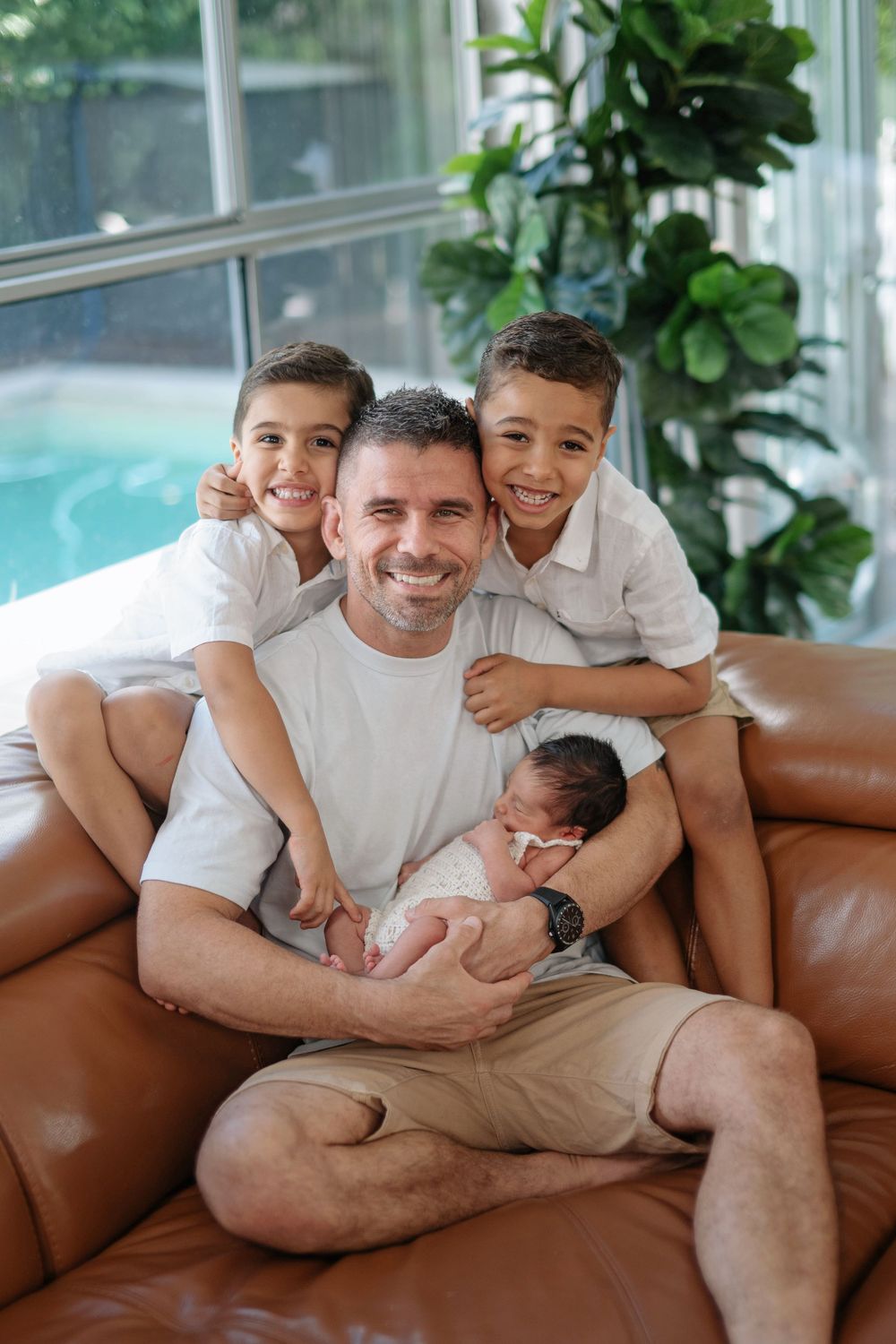 Happy father with two young sons and newborn baby on leather sofa by pool.