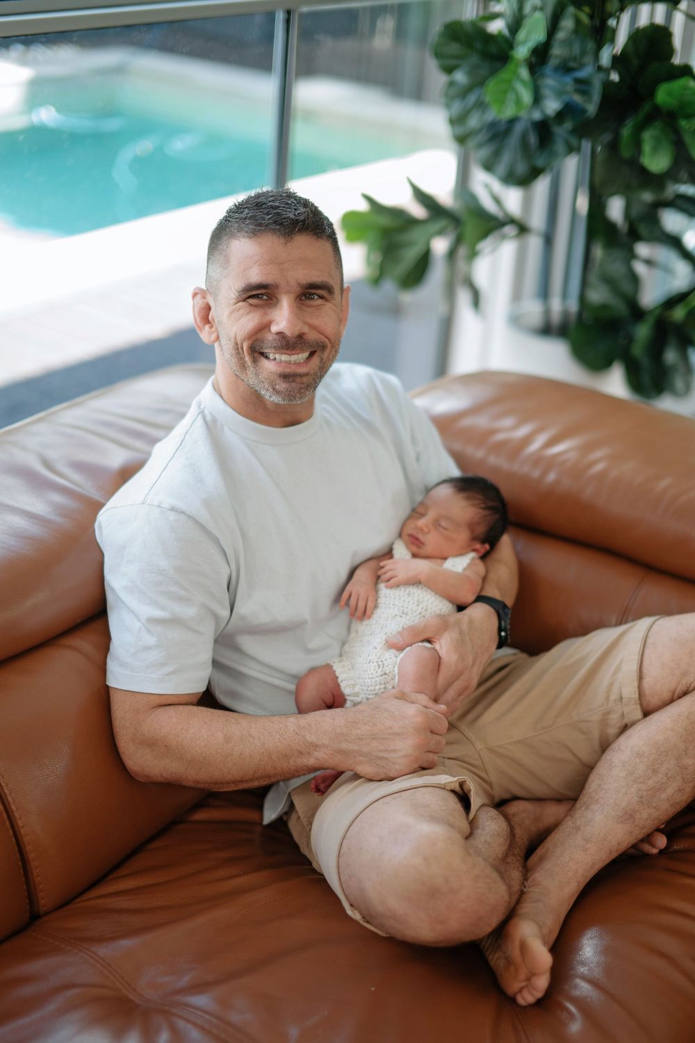 Smiling father holding sleeping newborn baby on brown leather sofa near indoor pool.
