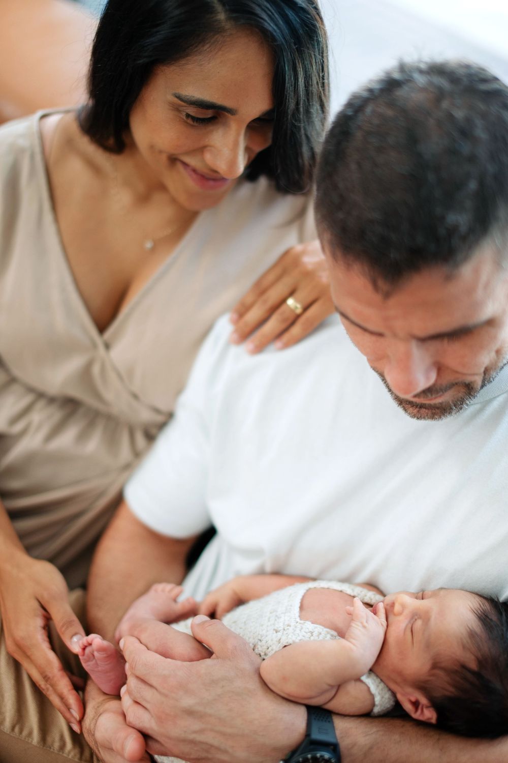 A couple tenderly gazes at their sleeping newborn baby, with the mother wearing a gold ring and the father in a white shirt.