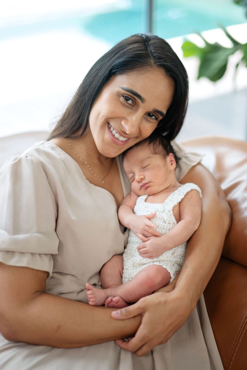 Smiling mother in beige dress holding sleeping newborn baby on leather sofa.