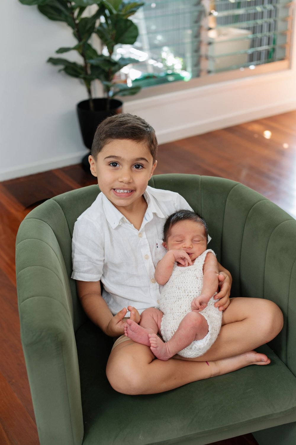 Young boy sitting in green velvet chair holding sleeping newborn sibling.