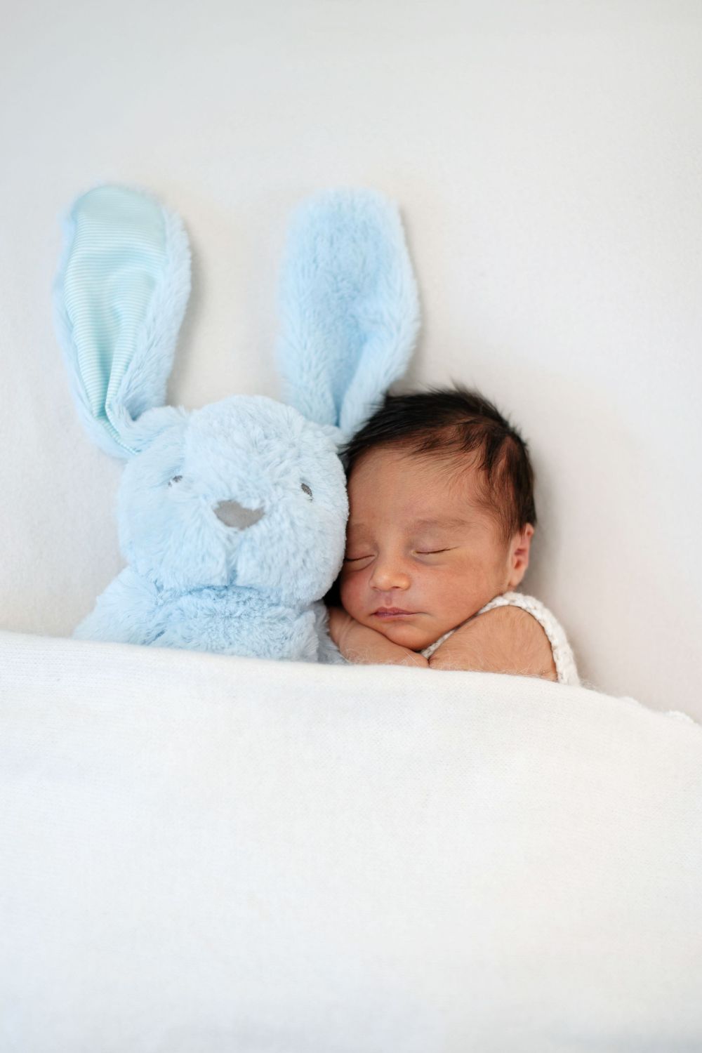 Newborn baby sleeping peacefully next to a soft blue stuffed bunny rabbit toy on a white blanket.