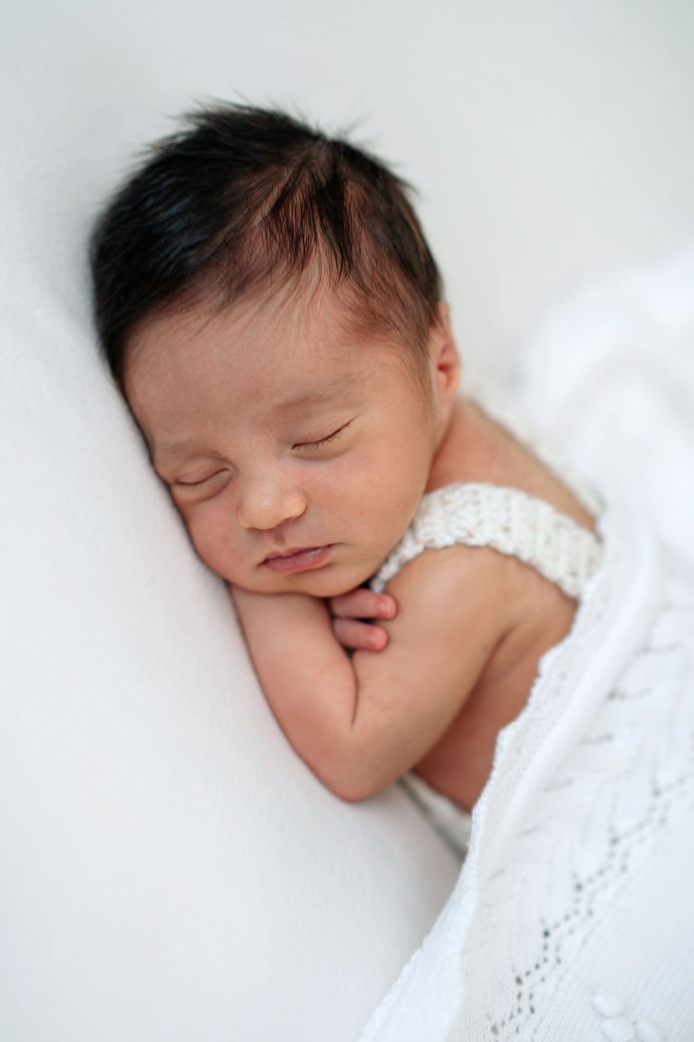 Peaceful newborn baby sleeping on white blanket with hands tucked under chin, dark hair, soft natural light.