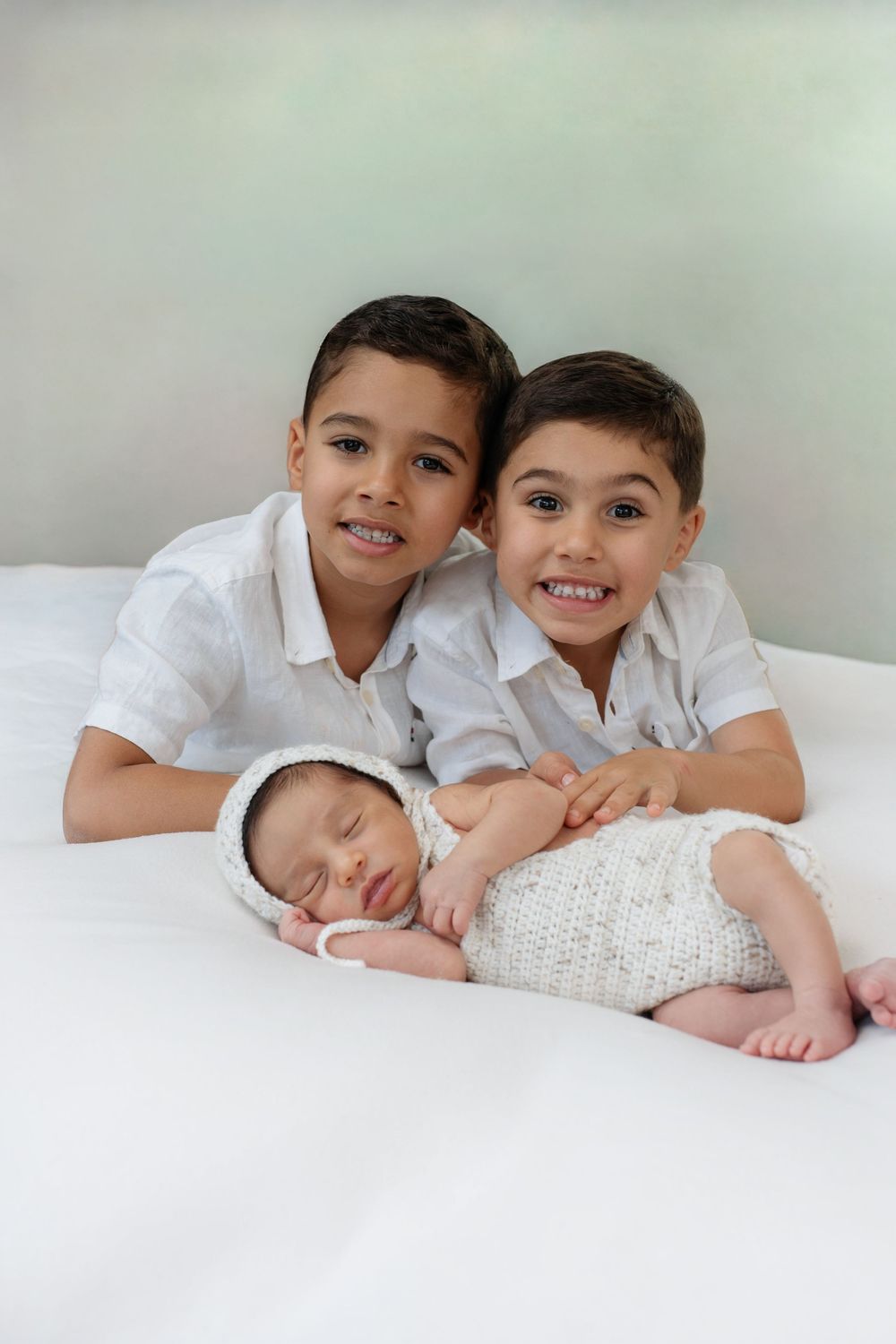 Two smiling boys in white shirts pose with a sleeping newborn baby wrapped in a knit outfit on a white bed.