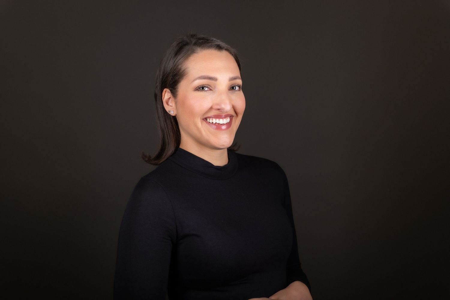 Smiling woman with dark hair in a black turtleneck poses confidently against a dark studio background.