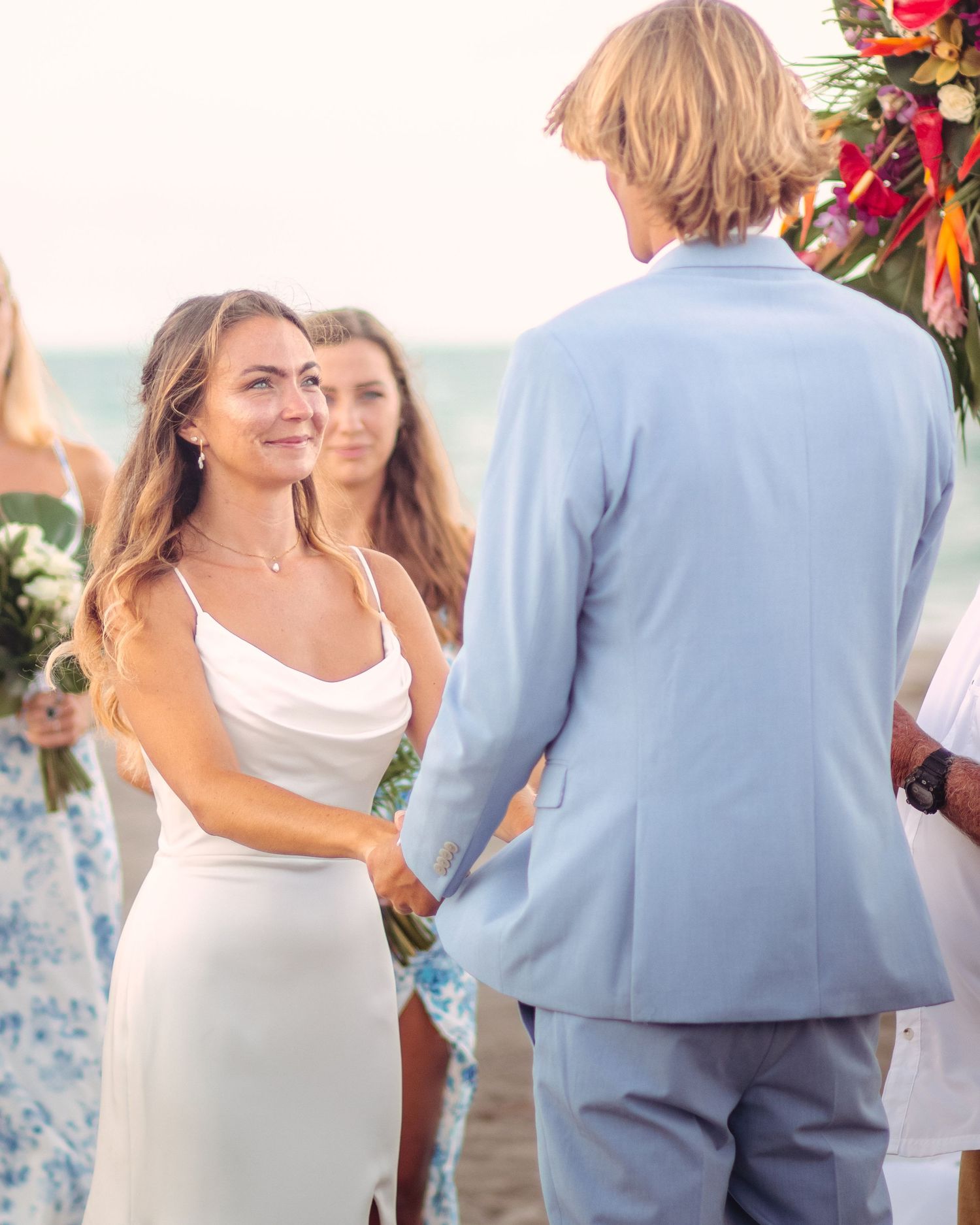 Bride in white dress holds hands with groom in light blue suit during outdoor beach wedding ceremony.