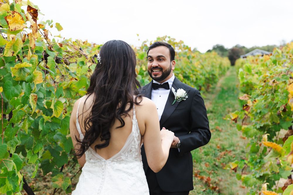 Groom in black tuxedo smiles at bride in white wedding gown among lush vineyard vines.