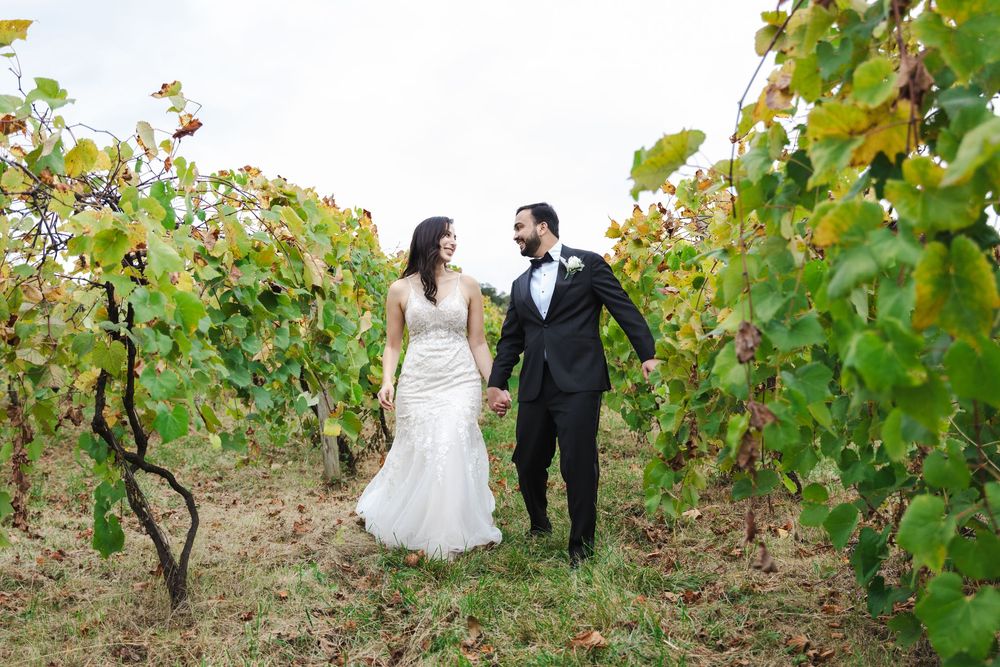 Bride and groom hold hands walking through lush vineyard rows in wedding attire on their special day.