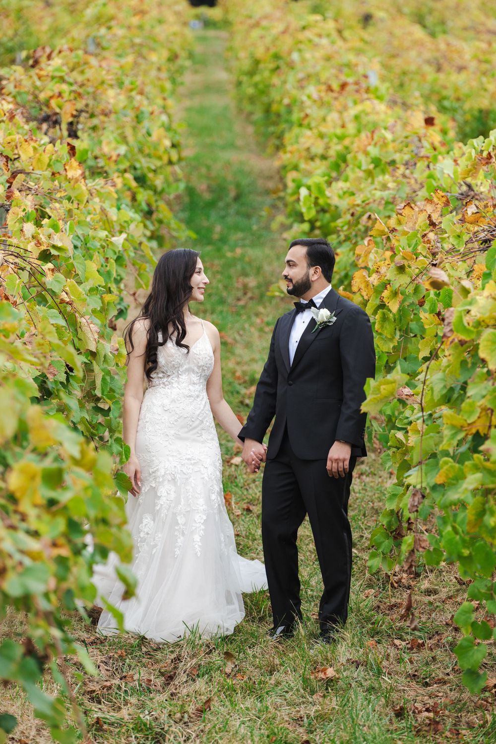 Bride and groom hold hands walking through lush vineyard rows in wedding attire, sharing a romantic glance.