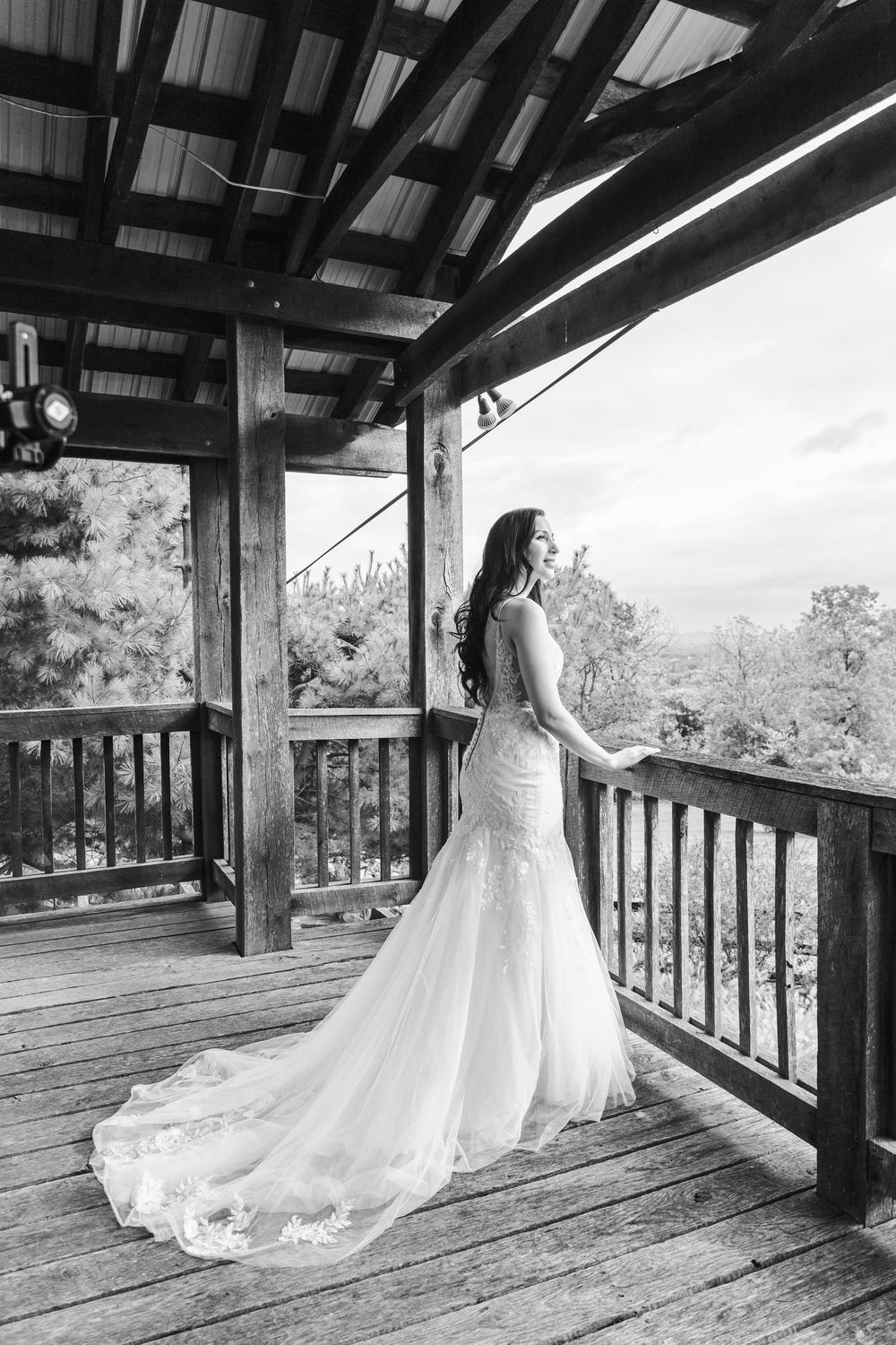 Bride in lace mermaid wedding gown standing on rustic wooden deck overlooking scenic forest view.