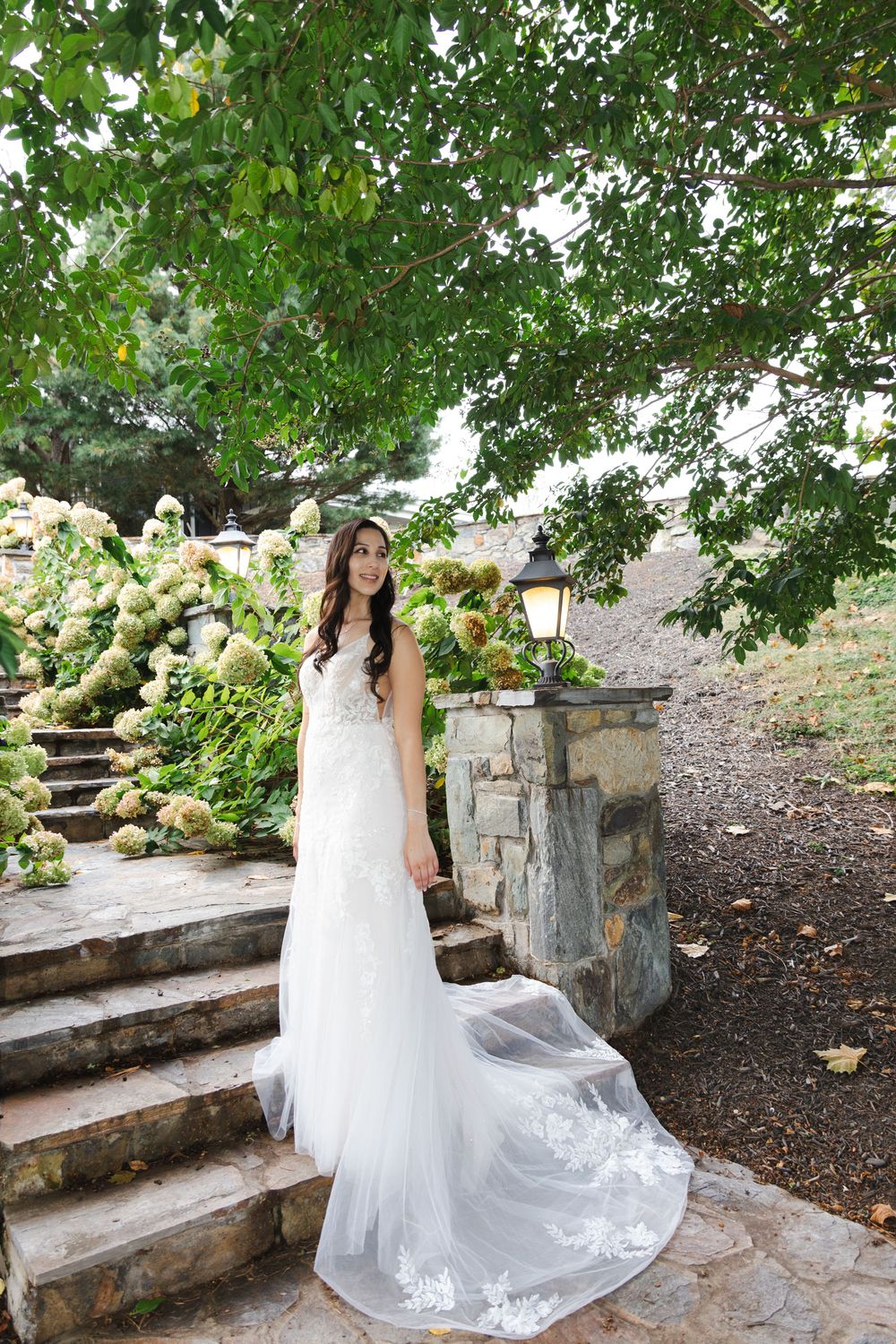 Bride in elegant lace wedding gown with train standing on stone steps amid hydrangea blooms.