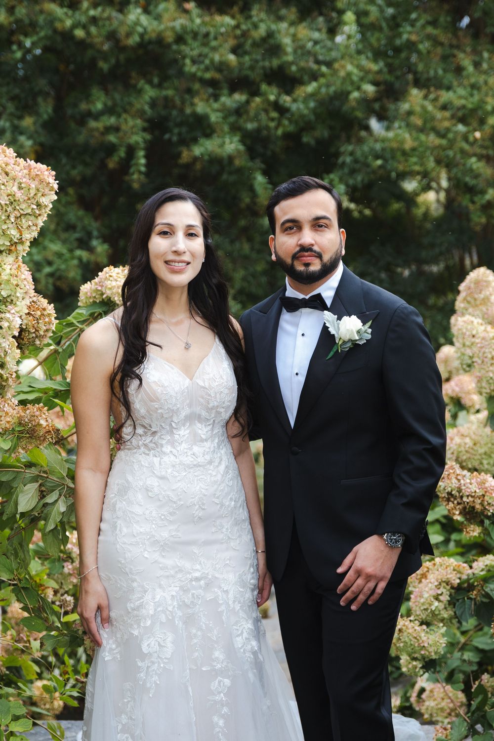 Bride and groom posing together outdoors among lush hydrangea flowers on their wedding day.