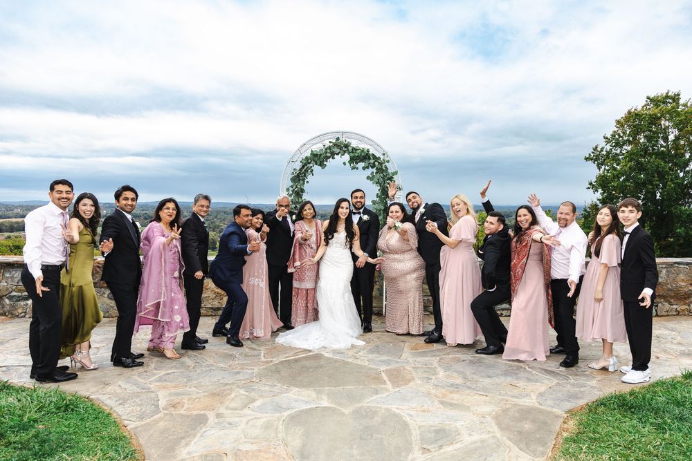 Wedding party poses together outdoors on stone patio with floral arch and scenic countryside views.