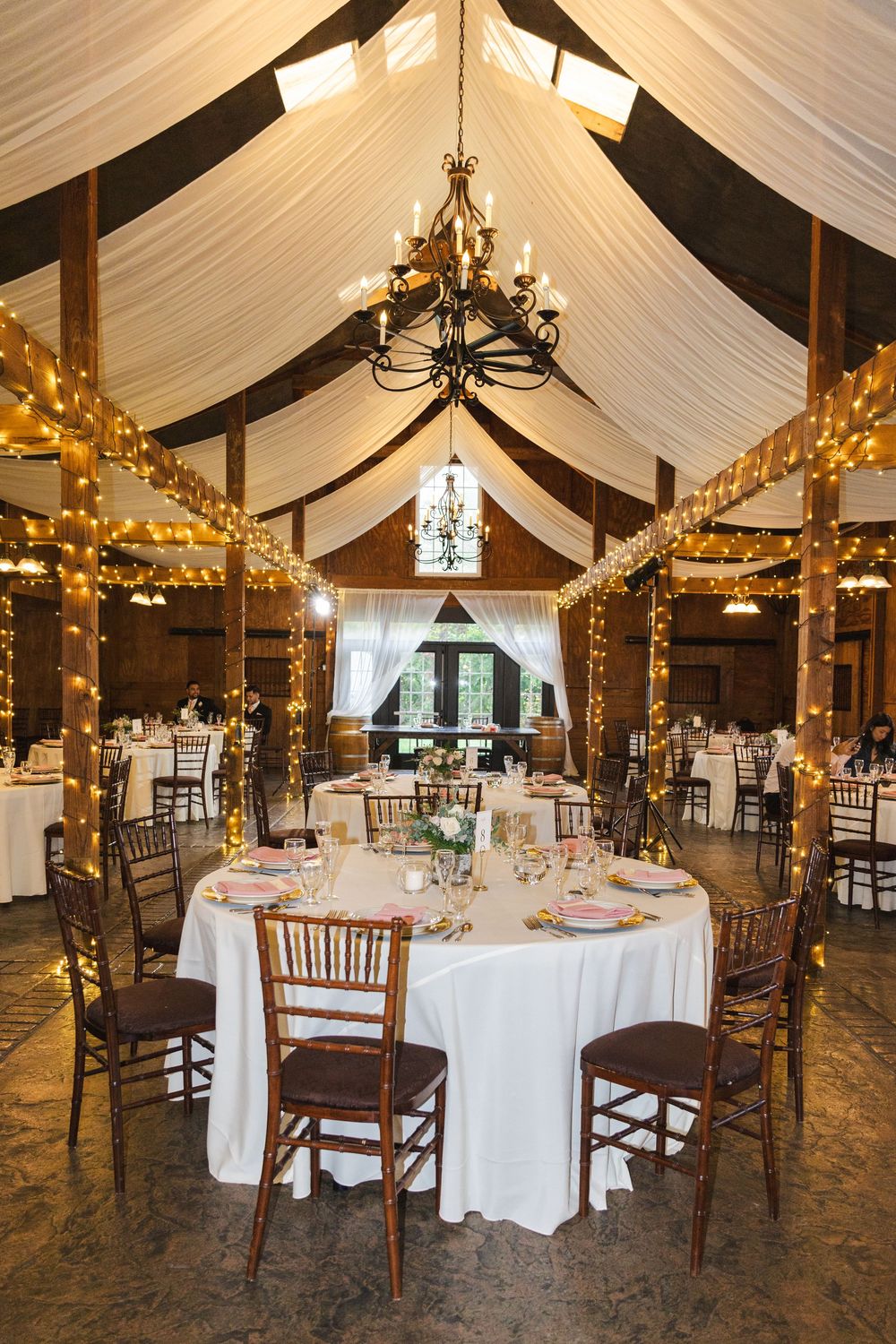 Elegant barn wedding reception with string lights, draped ceiling, and round tables.
