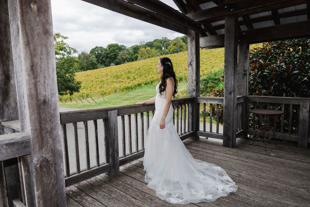 Bride in white gown with train stands on rustic wooden deck overlooking lush vineyard landscape.