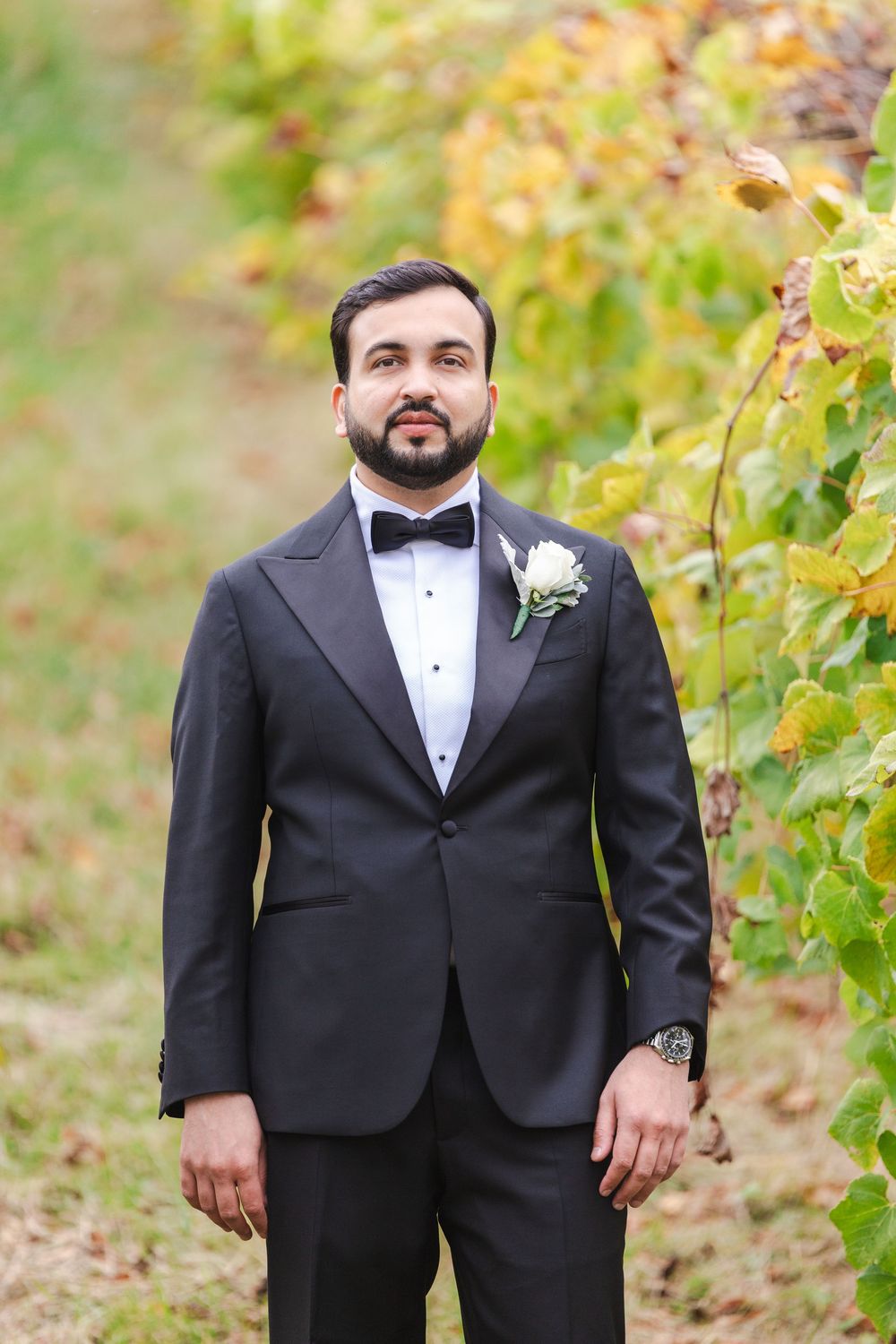 Groom in black tuxedo with bow tie and boutonniere stands among autumn vineyard rows.