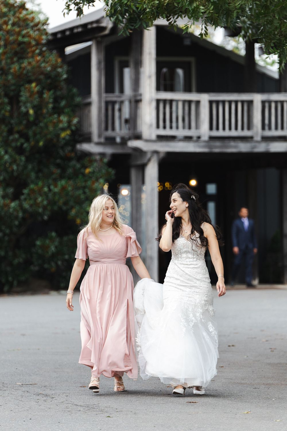 Bride in white gown walks with bridesmaid in pink dress outside rustic venue.
