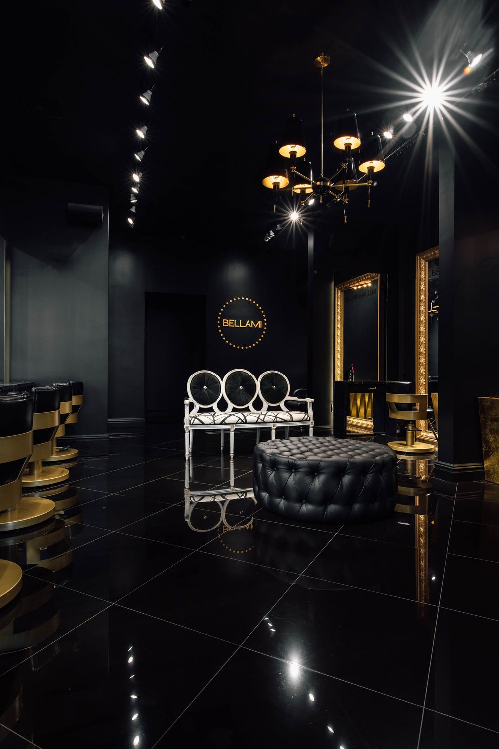 Luxurious black and gold hair salon interior with chandelier, tufted ottoman, and BELLAMI branding.