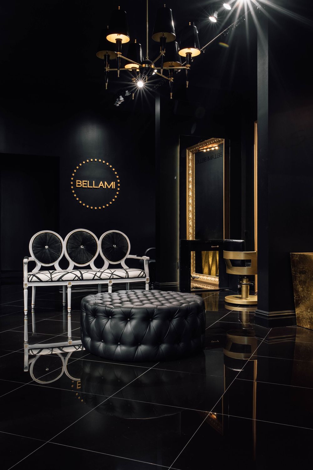 Bellami luxury salon interior with black tufted ottoman, ornate white bench, gold chandelier, and dark walls.
