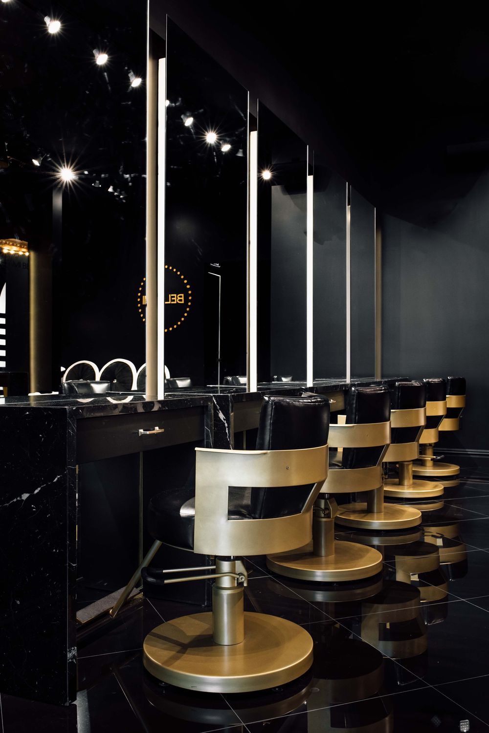 Luxury hair salon interior with gold styling chairs, black marble floors, and illuminated mirrors in an elegant dark setting.