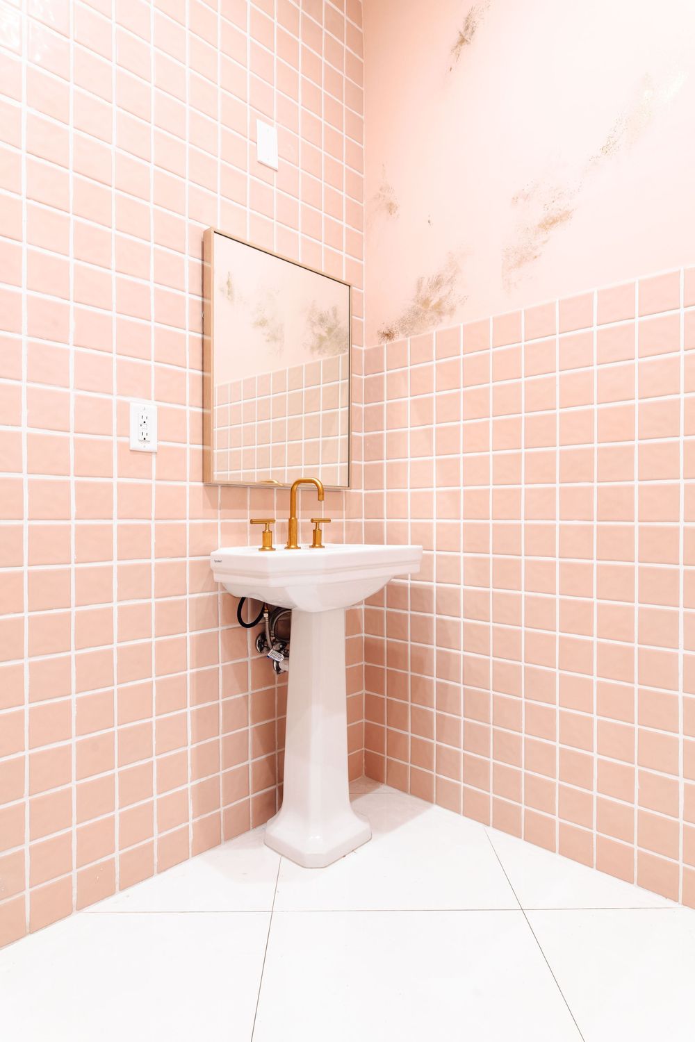 Elegant pink tiled bathroom corner featuring a white pedestal sink with gold faucet and small framed mirror.