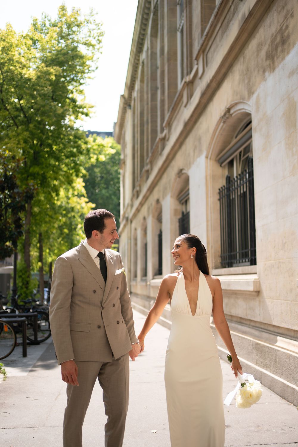 Bride and groom holding hands walking past a grand stone building on a sunny day, she in a white gown, paris wedding photographer civil
