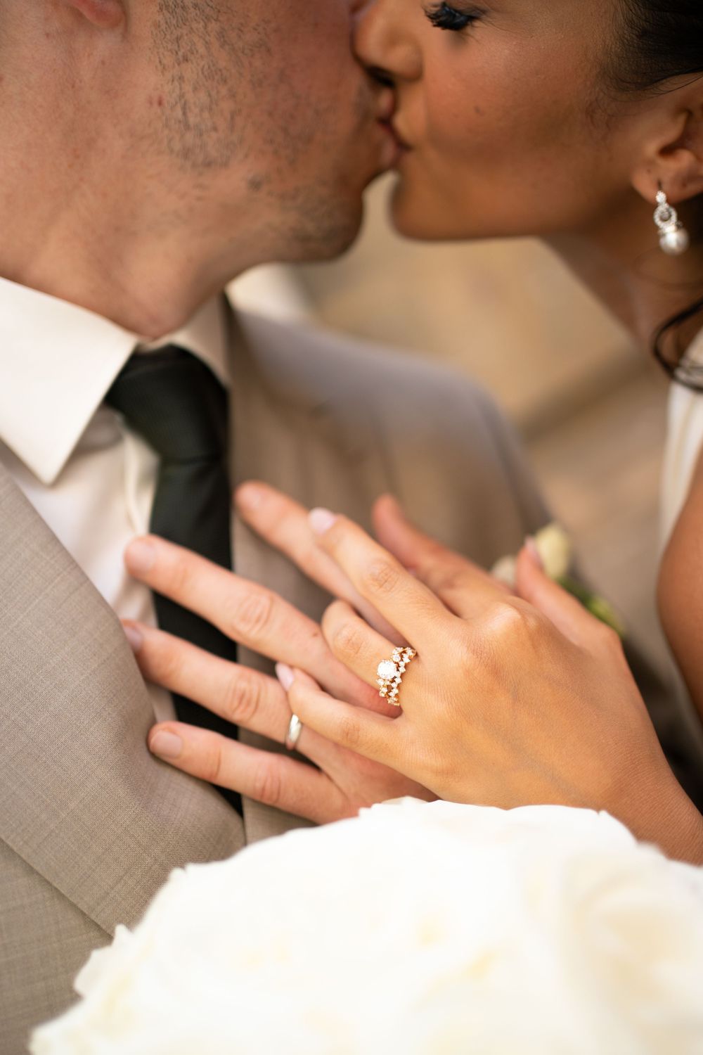 Newlywed couple sharing a kiss while bride displays her gold diamond wedding ring on groom's chest at wedding ceremony.