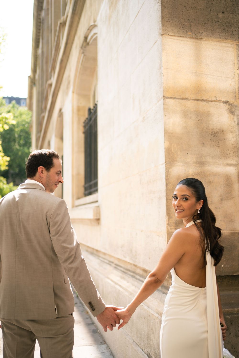 Bride in backless white gown holds hands with groom in tan suit while walking past a stone building. Paris film wedding photographer