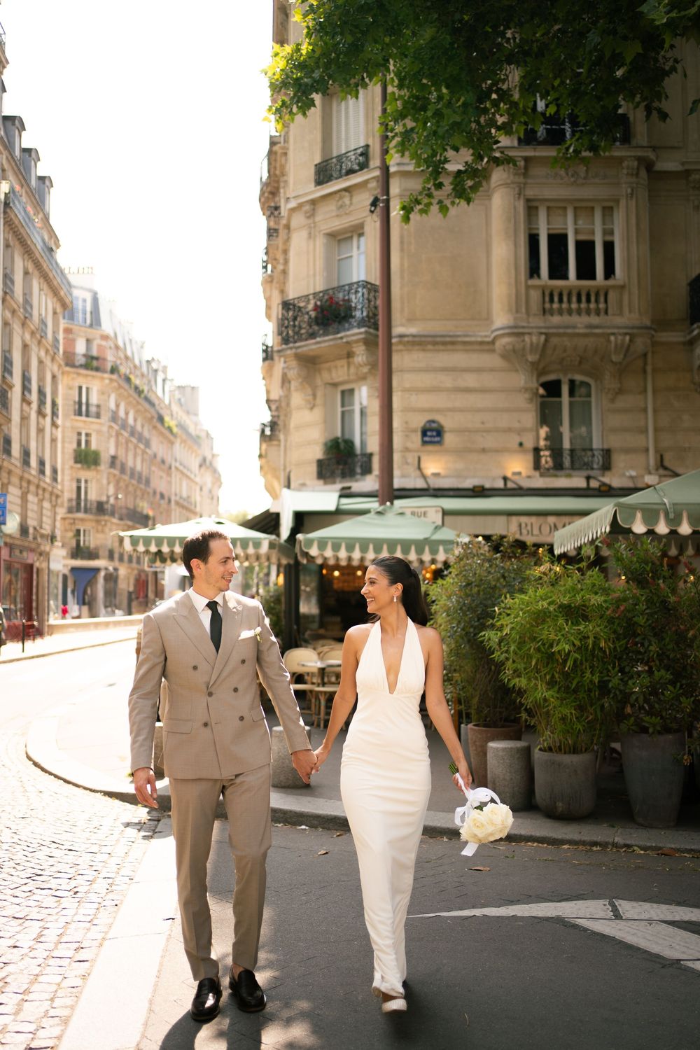 Bride and groom walk hand-in-hand on a sunny Paris street, she in a white gown, he in a beige suit. Candid paris wedding photography, film