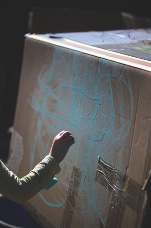 A child's hand drawing a blue chalk figure on a cardboard box in natural sunlight.