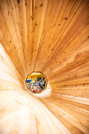 Interior view of a wooden tunnel with octagonal opening revealing workshop equipment beyond warm cedar planks.