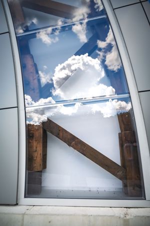 Modern glass window reflecting blue sky and white clouds with rustic wooden beams forming an X shape inside.