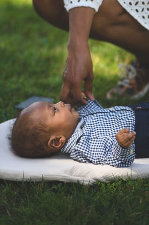 Adorable Black baby lying on a cushion in grass, looking up at a parent's hand in a checkered shirt.