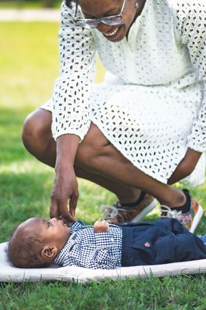 A smiling woman in a white lace dress crouches over a baby lying on a blanket in a sunny park.