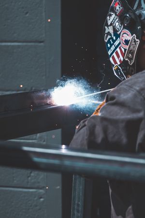 A welder in a sticker-covered helmet creates bright sparks while welding metal beams in a dark workshop.