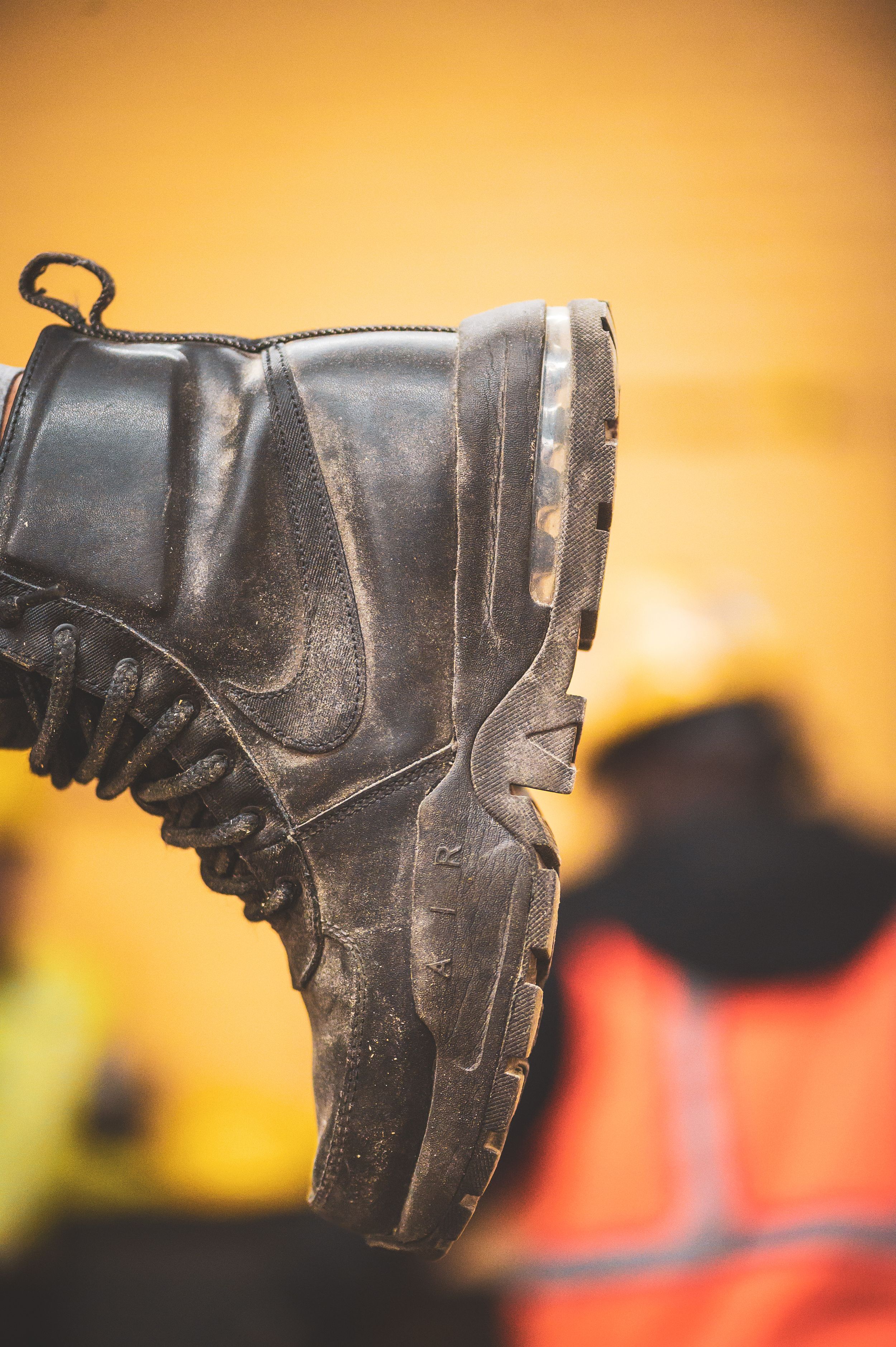 A worn black leather work boot held up against a blurred yellow background with a worker in safety gear visible.