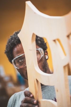 Craftsman wearing safety glasses inspecting a wooden chair frame in a woodworking workshop.
