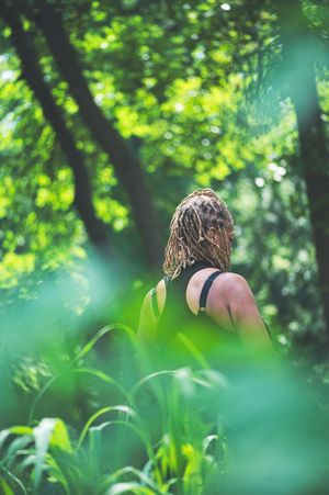 Person with dreadlocks in black top standing among lush green forest foliage in dappled sunlight.