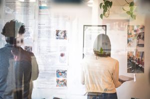 Two people viewing informational posters on a wall, photographed through glass in a modern office setting.