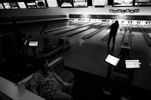 Black and white photo of people bowling at a retro bowling alley with glowing score screens.