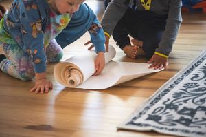 Two children unrolling a large roll of white paper on a wooden floor.