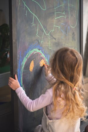 Young girl drawing a colorful smiley face on a black chalkboard with chalk.