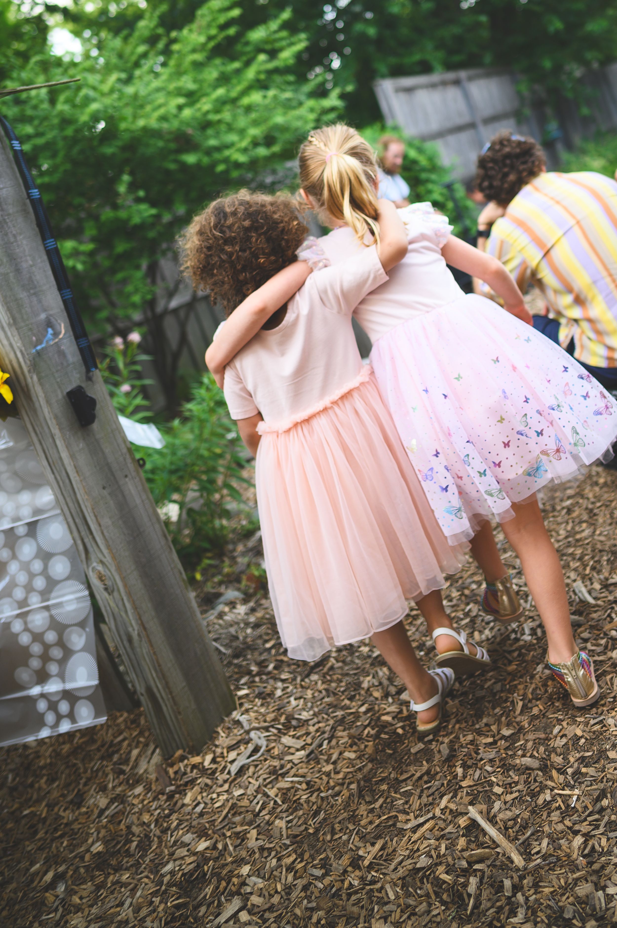 Two young girls in pink tulle dresses walking arm in arm through a garden.