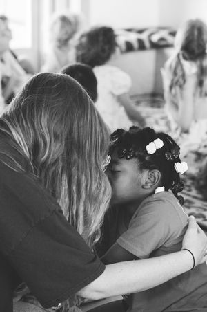 Black and white photo of a woman hugging a young girl with flower hair clips at an indoor gathering.