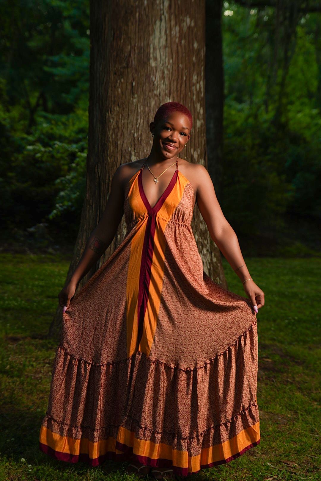 A smiling woman in a flowing orange and maroon maxi dress stands barefoot on grass in front of a large tree trunk.