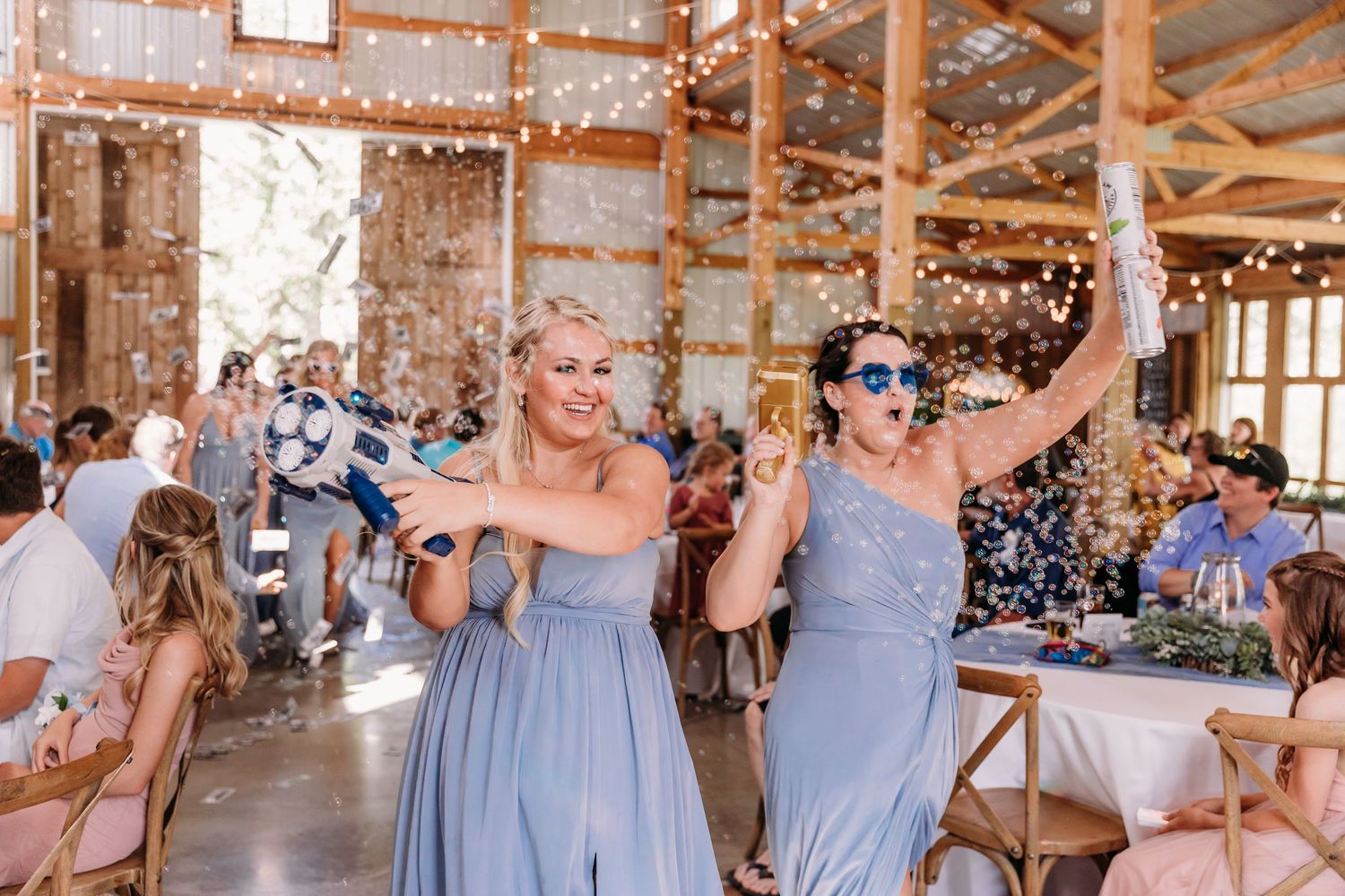 Two bridesmaids in blue dresses celebrate with confetti at a rustic barn wedding reception with string lights.