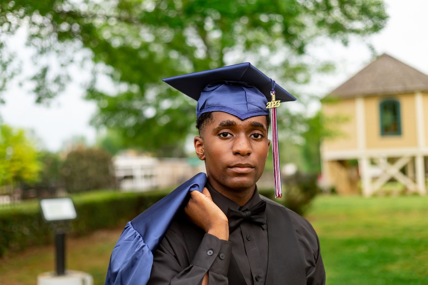 Young Black graduate in cap and gown poses outdoors, holding blue robe over shoulder with trees behind.